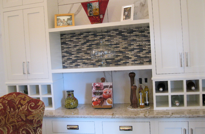 A kitchen counter with white cabinets, a patterned backsplash, a glass cake stand, a jar of olives, a cookbook, and spice grinders.